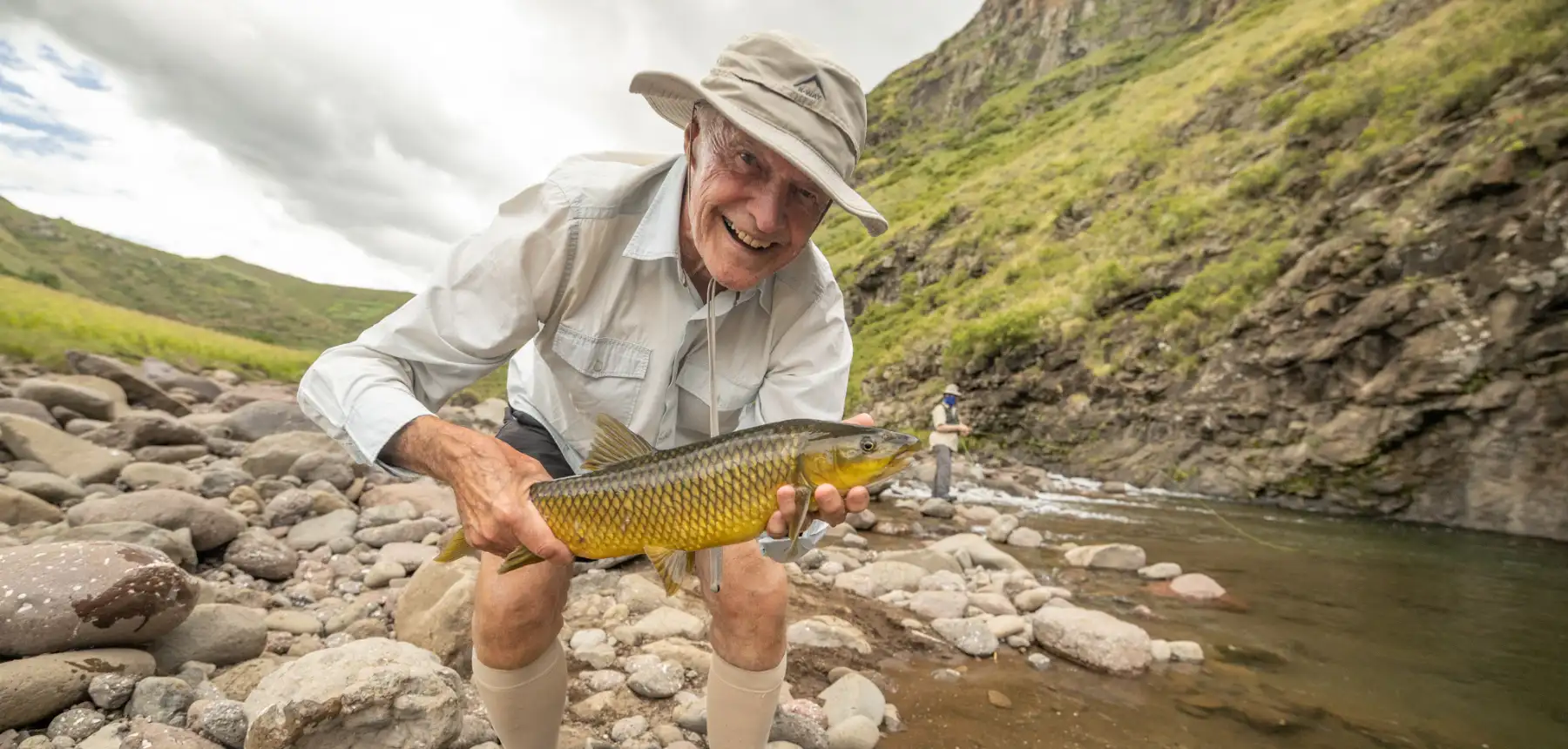 Man holding yellowfish beside rocky river in outdoor setting